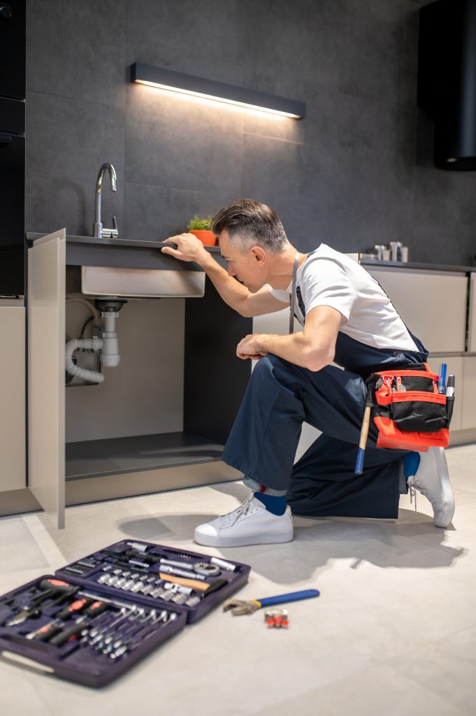 Man Kneeling Down Touching Sink Inspecting Pipes 1